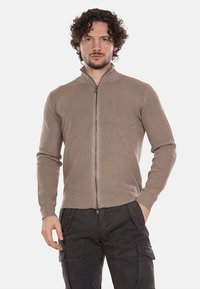Cardigan beige a costine con zip alta, caratterizzato da una zip argentata e maniche aderenti. Indossato sopra pantaloni cargo scuri.