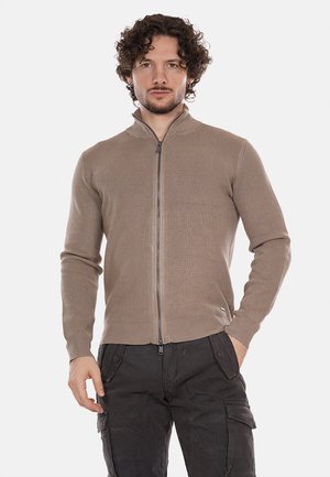 Cardigan beige a costine con zip alta, caratterizzato da una zip argentata e maniche aderenti. Indossato sopra pantaloni cargo scuri.