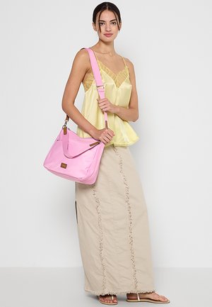 Femme portant un débardeur en satin jaune, une longue jupe beige, des sandales beiges, portant un grand sac à bandoulière rose avec une large sangle sur fond blanc.
