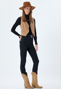 Chaleco marrón con botones sobre un suéter de cuello alto negro, combinado con jeans negros de cintura alta y botas vaqueras color beige con bordados.