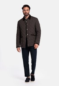 Gequilted bruine jas met een donkere kraag, voorzakken en drukknoopsluitingen. Draagt over een grijs shirt, gecombineerd met donkere broek en zwarte instappers.