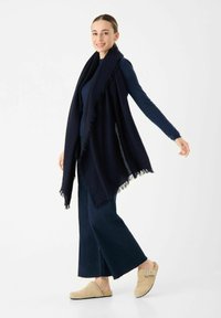 Marineblauwe wikkel sjaal met franjes, gedragen over een donkere top en wijde jeans, gecombineerd met beige klompen. Gladde textuur, ontspannen pasvorm.
