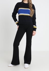 Granatowy sweter w niebieskie, białe i żółte poziome paski; zestawiony z czarnymi spodniami dzwonami i białymi trampkami.