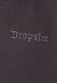 Sort stof med en tekstureret finish har ordet "Dropsize" broderet i en kontrasterende lys lilla gotisk skrifttype.