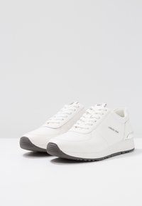 Sneaker bianca con tomaia in pelle liscia, chiusura con lacci, suola in gomma grigia e un sottile accento metallico sul tallone.