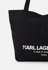 Černá plátěná taška s dlouhými uchy. Obsahuje bílý potisk textu: "KARL LAGERFELD 21 RUE ST-GUILLAUME PARIS." Jednoduchý design, pevný materiál.