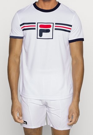 Homme portant une tenue de sport blanche avec un t-shirt à logo rayé bleu marine et rouge et un short blanc, debout devant un fond uni.