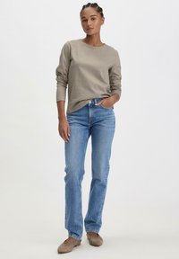 Graues Langarmshirt mit rundem Halsausschnitt, kombiniert mit hellblauen geraden Jeans. Das Modell trägt beigefarbene Slipper. Neutraler Hintergrund.
