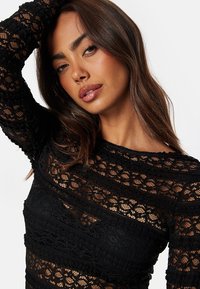 Top in pizzo nero con intricate fantasie floreali, maniche lunghe e scollatura a barca, caratterizzato da una trama semi-trasparente e una silhouette aderente.