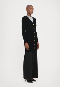 Black ribbed cardigan with long sleeves, gold buttons down the front, layered over a light gray top, paired with wide black trousers and heels.