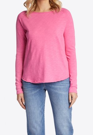 Kobieta ubrana w różową koszulę z długim rękawem i jasnoniebieskie dżinsy, stojąca na białym, gładkim tle.