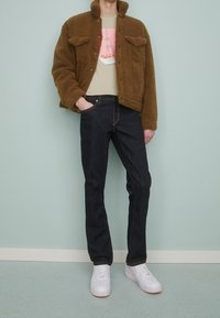 Personne portant une veste en peluche marron, un t-shirt graphique beige, un jean bleu foncé et des baskets blanches, debout contre un mur bleu clair.