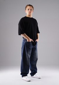 Young man with tattooed arm wearing black oversized T-shirt, loose blue jeans, white sneakers, standing with hands in pockets on gray background.