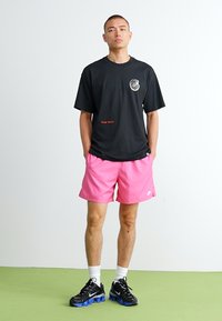 Camiseta negra con un logo redondo y texto rojo, combinada con pantalones cortos rosas. El modelo lleva zapatillas negras con una suela azul y calcetines blancos.