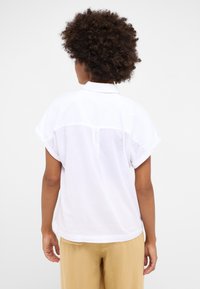 Camisa blanca de manga corta con cuello y diseño de yugo trasero. Tejido suave y ligero que se drapea de forma holgada. Detalle de costura visible a lo largo de las mangas.