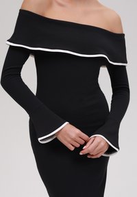 Vrouw in een zwarte off-shoulder jurk met witte bies op de kraag en uitlopende mouwen, handen voorzichtig op de taille gekruist.