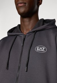 Pilkas zip-up džemperis, pagamintas iš tekstūruoto audinio, su juodu užtrauktuku ir raišteliais, ant krūtinės su baltu EA7 Emporio Armani logotipu.