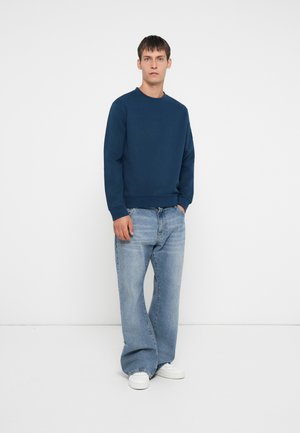 Marinblå sweatshirt med rund halsringning, matchad med ljusblå, löst sittande jeans. Modellen står mot en vit bakgrund.