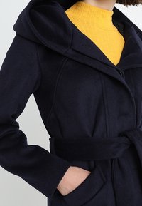 Cappotto in lana blu navy con cappuccio ampio, vita con cintura e tasche laterali, caratterizzato da una texture liscia e linee pulite.