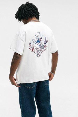 Homme aux cheveux noirs bouclés portant un t-shirt blanc avec un graphique coloré de cowboy sur un cheval et un jean bleu, se tenant contre un fond neutre.
