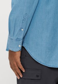 Jasnoniebieska koszula jeansowa z długimi rękawami, wyposażona w mankiety na guziki i zaokrąglony dół. Zbliżenie na rękaw i dłoń ukazuje teksturę tkaniny.