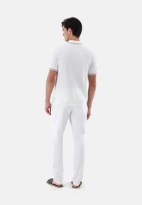 Weißes Polohemd mit gestreiften Kragen, kombiniert mit weißen Hosen. Der Stoff scheint leicht und mit einer glatten Textur zu sein. Sichtbares lässiges Schuhwerk.
