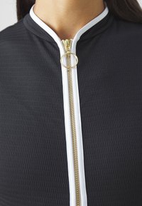 Schwarze strukturierte Zip-Jacke mit weißem Besatz und goldener Reißverschluss mit rundem Ziehgriff. Glattes Material mit modernem Design.