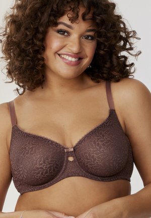 Femme aux cheveux bouclés souriante, portant un soutien-gorge marron en dentelle avec armatures, posant devant un fond clair uni.