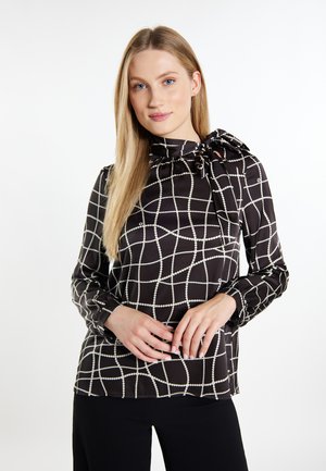 Frau mit langen blonden Haaren, die eine schwarze Bluse mit weißem Gittermuster und einer Krawatte trägt, steht mit vor dem Körper verschränkten Händen.
