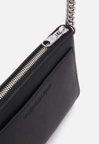 Pochette nera in ecopelle vegana con cerniera argentata, caratterizzata da una superficie testurizzata, logo in rilievo e tracolla a catena metallica.