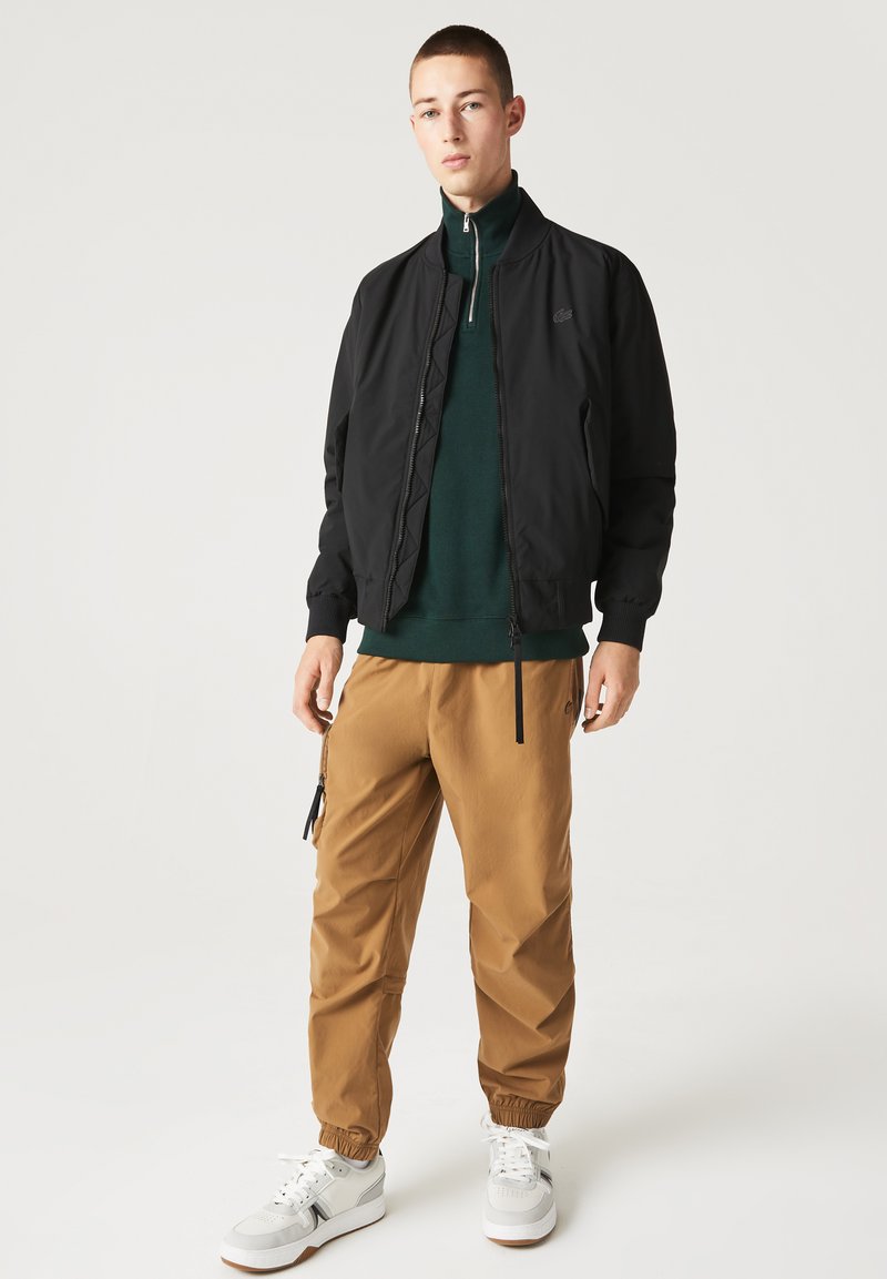 Veste bomber noire avec une fermeture éclair sur le devant, superposée à un pull zippé vert foncé. Pantalons à poignets élastiques beiges et baskets blanches complètent la tenue.