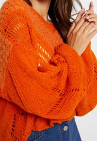 Jasnopomarańczowy sweter z grubym splotem, z otwartymi wzorami dzianiny, szerokimi rękawami i luźnym krojem.