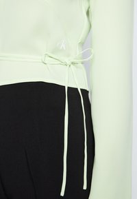 Top de mangas longas verde claro com um design cruzado, apresentando uma cintura amarrada e detalhe de logotipo branco, combinado com parte de baixo pretas. Textura suave.