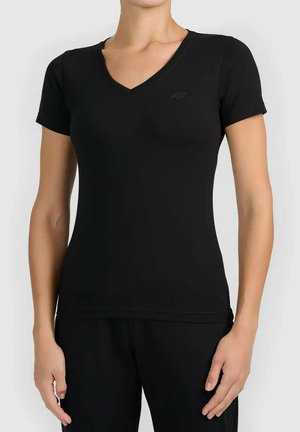 Kobieta ubrana w dopasowaną czarną koszulkę z krótkim rękawem i dekoltem w serek oraz czarne spodnie, stojąca na jednolitym tle.