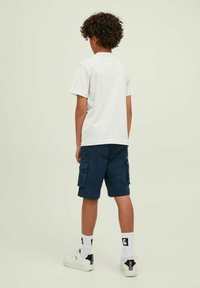 T-shirt bianca, pantaloni cargo blu navy con tasche, calze bianche con logo e sneakers bianchi. Tessuto strutturato e vestibilità rilassata.