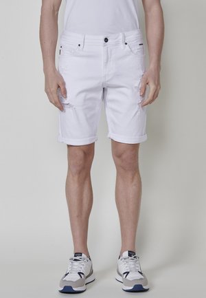 Bermudas de mezclilla blanca con detalles desgastados, dobladillos enrollados, cinco bolsillos y herrajes negros. Llevadas con zapatillas gris y azul marino.