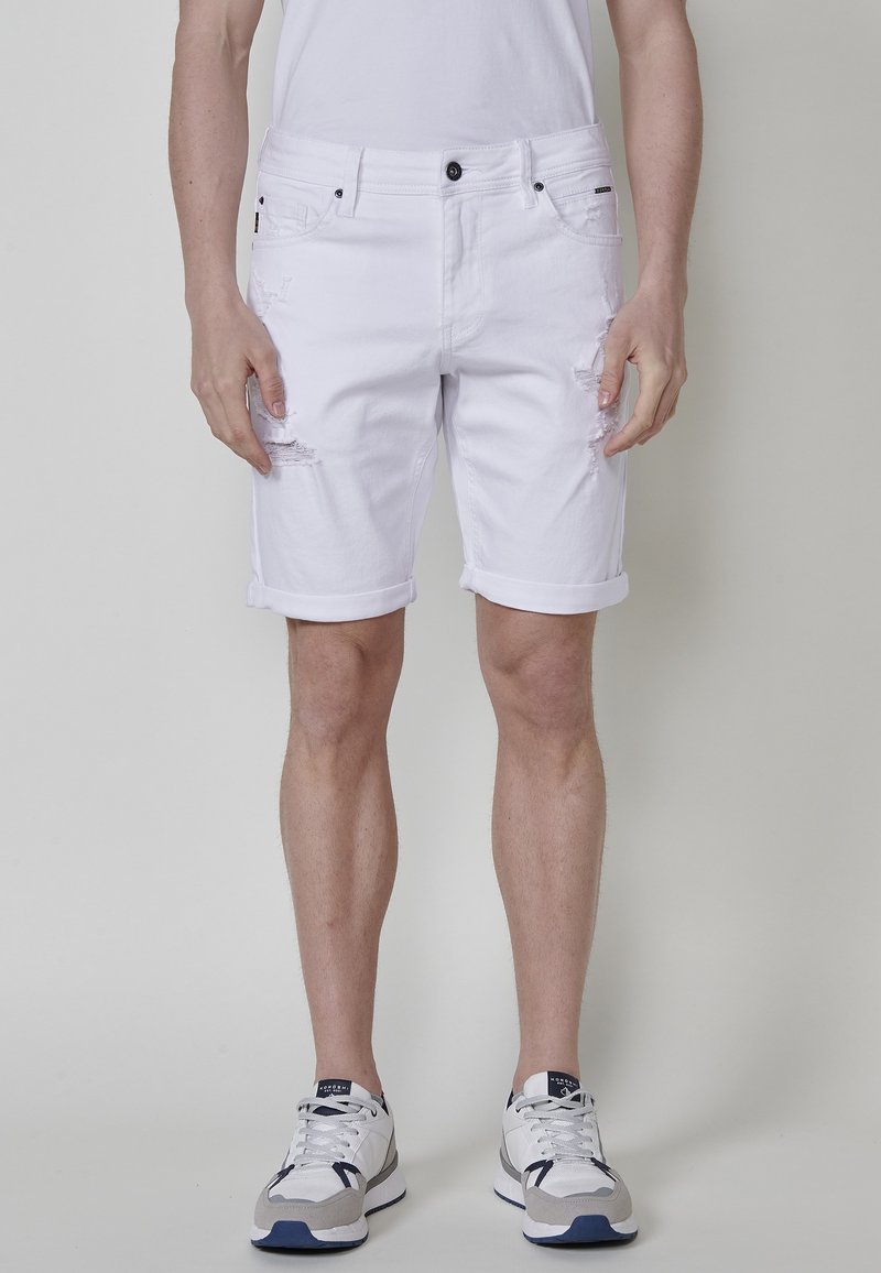 Bermudas de mezclilla blanca con detalles desgastados, dobladillos enrollados, cinco bolsillos y herrajes negros. Llevadas con zapatillas gris y azul marino.