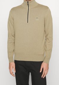 Béžový svetr na zip s vysokým límcem, žebrovanými manžetami a lemem. Na hrudi má malou čtvercovou logo. Tkanina má hladkou strukturu.