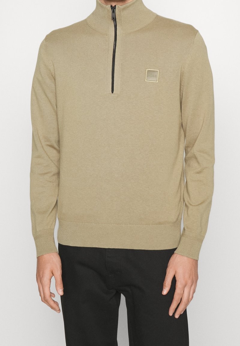 Béžový svetr na zip s vysokým límcem, žebrovanými manžetami a lemem. Na hrudi má malou čtvercovou logo. Tkanina má hladkou strukturu.