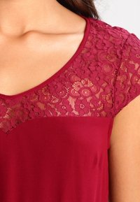 Top burdeos con cuello redondeado, cortas mangas de encaje y superposición de encaje floral en la zona de los hombros, con un cuerpo de tela suave.