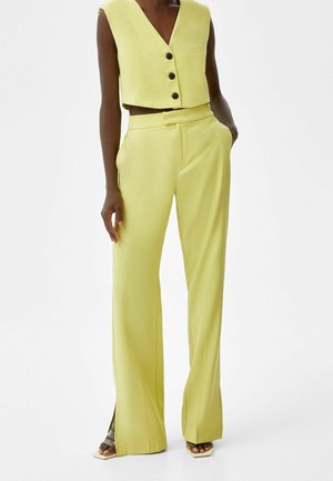 Gilet jaune court avec trois boutons et une poche, associé à un pantalon taille haute à jambes larges avec fentes latérales et texture lisse.