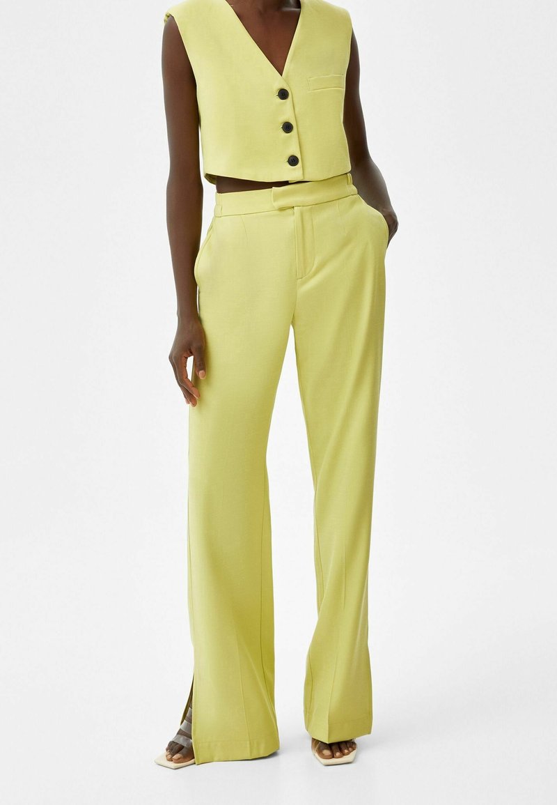 Gilet jaune court avec trois boutons et une poche, associé à un pantalon taille haute à jambes larges avec fentes latérales et texture lisse.