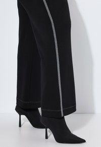 Nogi w czarnych szerokich spodniach z szarymi bocznymi paskami i czarnych szpiczastych skórzanych botkach na białej podłodze.