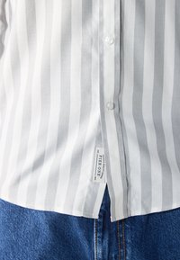 Wit en lichtgrijs gestreept shirt met een knoopsluiting en een label aan de onderlinkerzijde, gecombineerd met blauwe denim jeans.