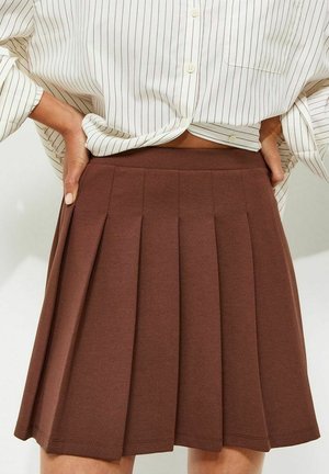 Vrouw draagt een bruine geplooide rok met hoge taille en een los wit hemd met dunne verticale strepen, ingestopt.