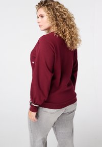 Vrouw met krullend blond haar, gekleed in een bordeauxrode longsleeve en lichtgrijze jeans, staande en naar de zijkant kijkend tegen een witte achtergrond.