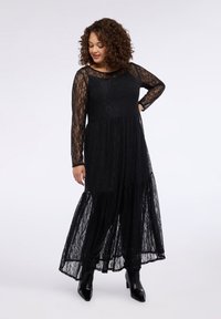 Czarna sukienka maxi z koronki z długimi rękawami, warstwową spódnicą i jednolitym gorsetem. Posiada dekolt w łódkę i teksturowany materiał. Połączona z czarnymi botkami za kostkę.