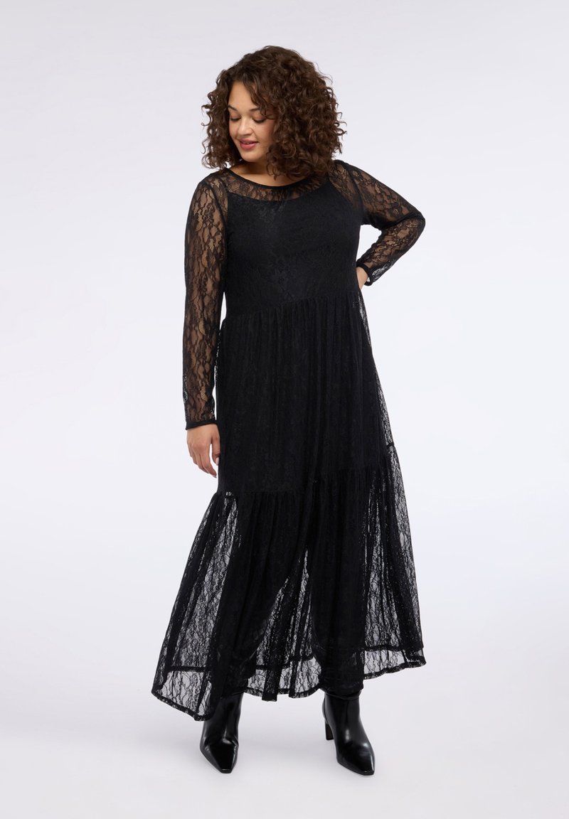 Czarna sukienka maxi z koronki z długimi rękawami, warstwową spódnicą i jednolitym gorsetem. Posiada dekolt w łódkę i teksturowany materiał. Połączona z czarnymi botkami za kostkę.