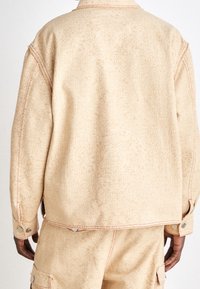 Beige denimjacka med en subtil mönstrad textur och synliga sömnadsdetaljer. Har knappstängningar med tryckknappar och en avslappnad passform.