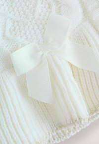 Weißer Strickstoff mit weicher Textur, der in der Mitte mit einer großen weißen Schleifenapplikation verziert ist. Subtile Rippstruktur und metallische Akzente.